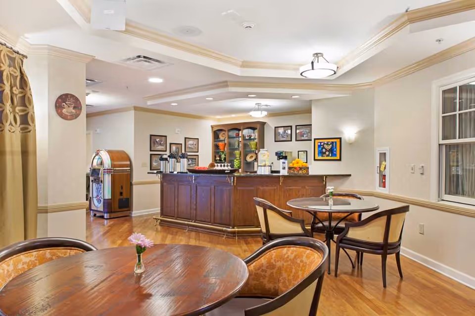 A cozy common area with wooden floors and round tables with cushioned chairs. In the background, there is a wooden counter with coffee dispensers, a fruit basket, and a small cabinet with decorative items. The room is softly lit with ceiling lights and wall sconces, and there is a jukebox against the far wall.