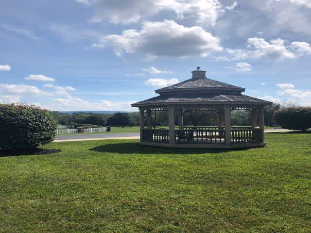 A wooden gazebo with a shingled roof situated on a well-maintained grassy area under a partly cloudy blue sky. There are bushes on either side of the gazebo and a paved path or road behind it, with trees and a distant landscape visible in the background.