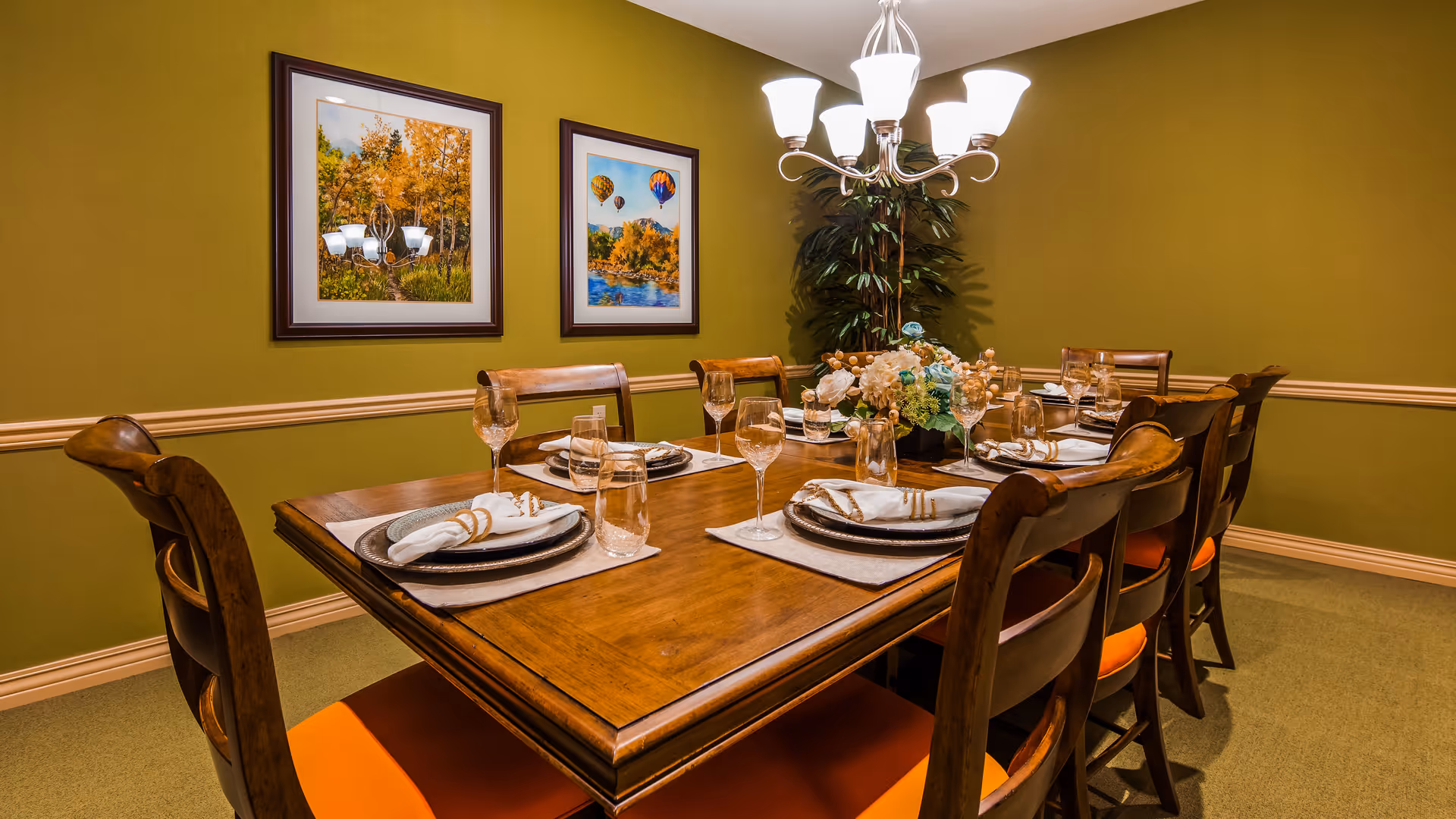 A dining room with a wooden table set for eight people. The table is arranged with plates, napkins, glasses, and a floral centerpiece. The room has green walls with two framed pictures and a chandelier overhead. There is a plant in the corner of the room.