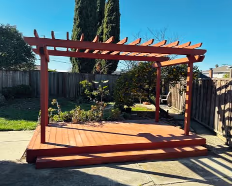 A wooden pergola structure with a slatted roof and steps leading up to the platform, situated in a backyard garden area with grass, plants, and a wooden fence under a clear blue sky.