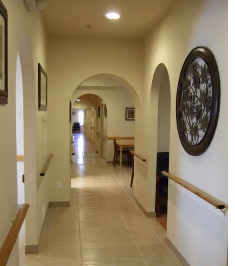 Well-lit interior hallway with tiled floor, arched doorways, wall-mounted handrails and a table visible at the far end.