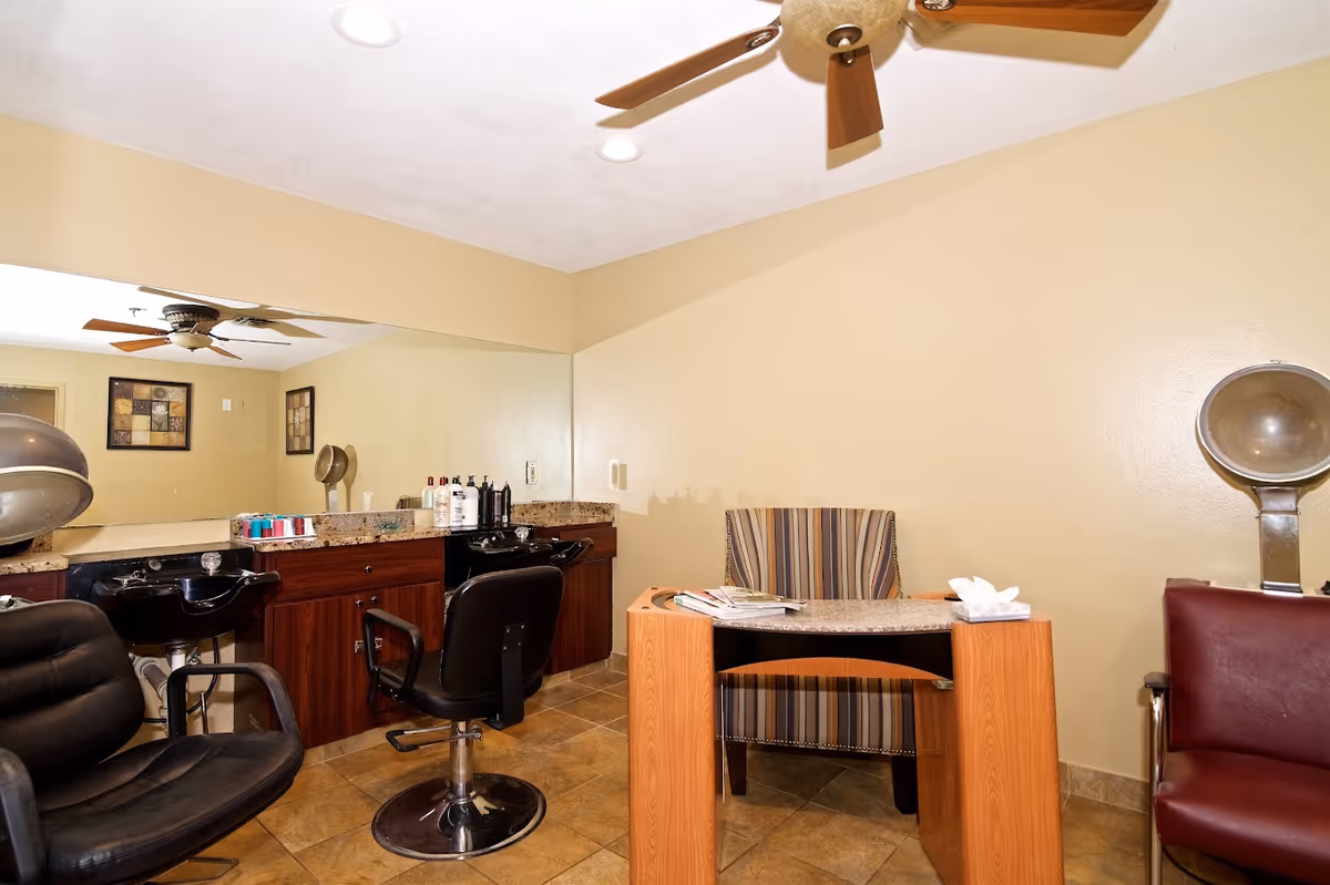 Interior view of a salon area in a senior living facility featuring two black salon chairs in front of a large mirror with hair washing sinks, a wooden manicure table with a striped chair, and a vintage hair dryer chair against a beige wall.