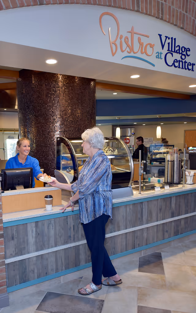 An elderly woman receives a pastry from a smiling staff member at a bistro counter inside a senior living facility.