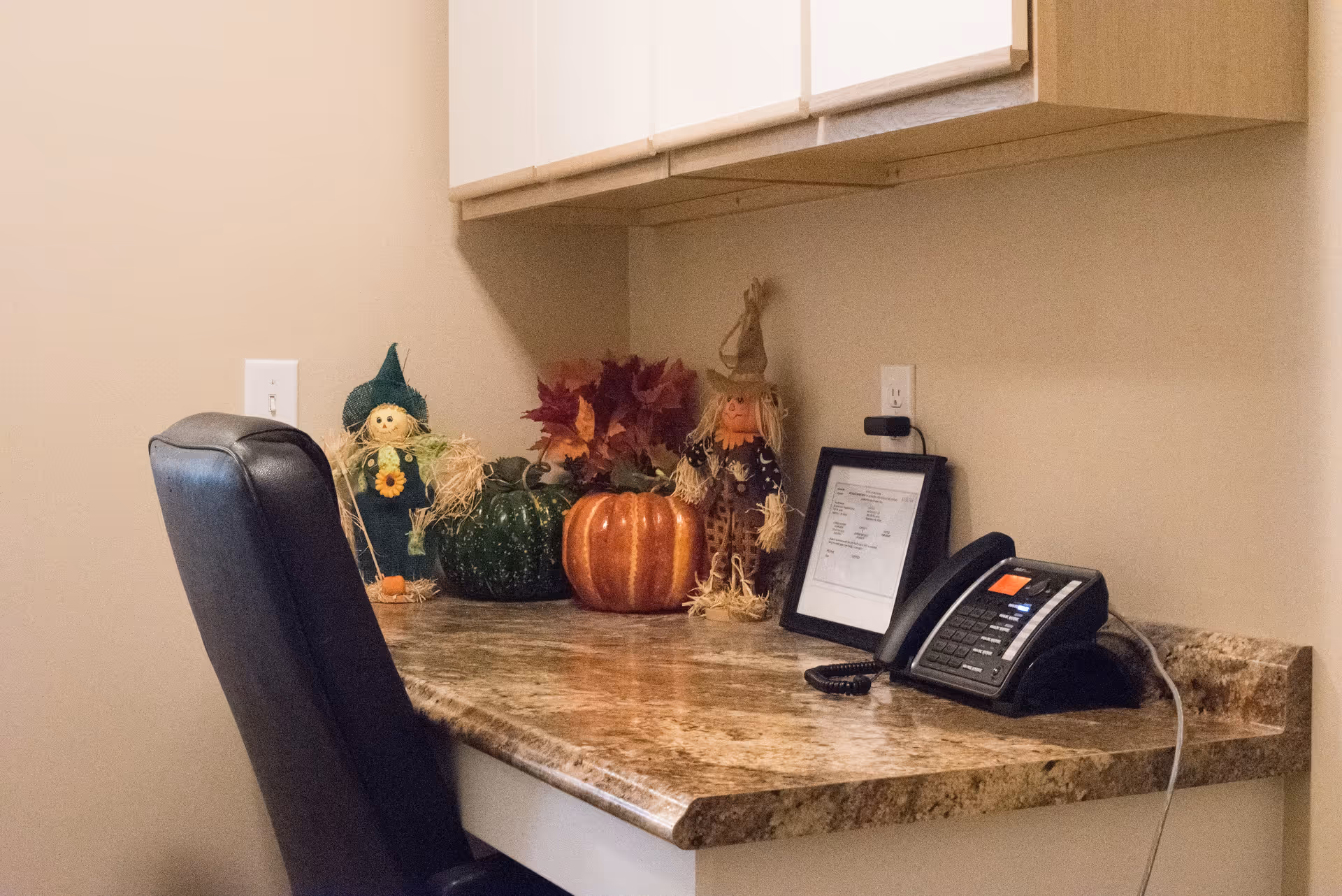 A small office or workspace with a marble-patterned desk, a black office chair, a telephone, a framed document, and autumn-themed decorations including two scarecrow figurines, a green gourd, an orange pumpkin, and artificial fall leaves.