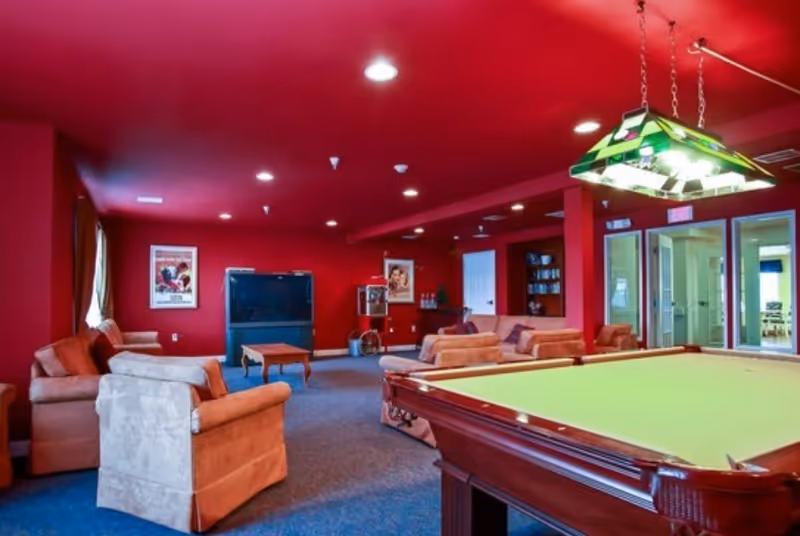 Recreation/lounge room with a pool table in the foreground, sofas and armchairs, and a TV against a red-painted wall.