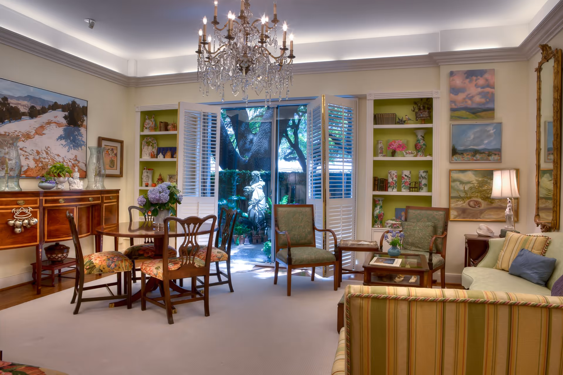 A cozy living room with a chandelier hanging from the ceiling, a round wooden dining table with four floral upholstered chairs, two green armchairs, a striped sofa, and a light green couch. The room has built-in shelves with decorative items and paintings on the walls. French doors with white shutters open to a garden area with a statue and trees.