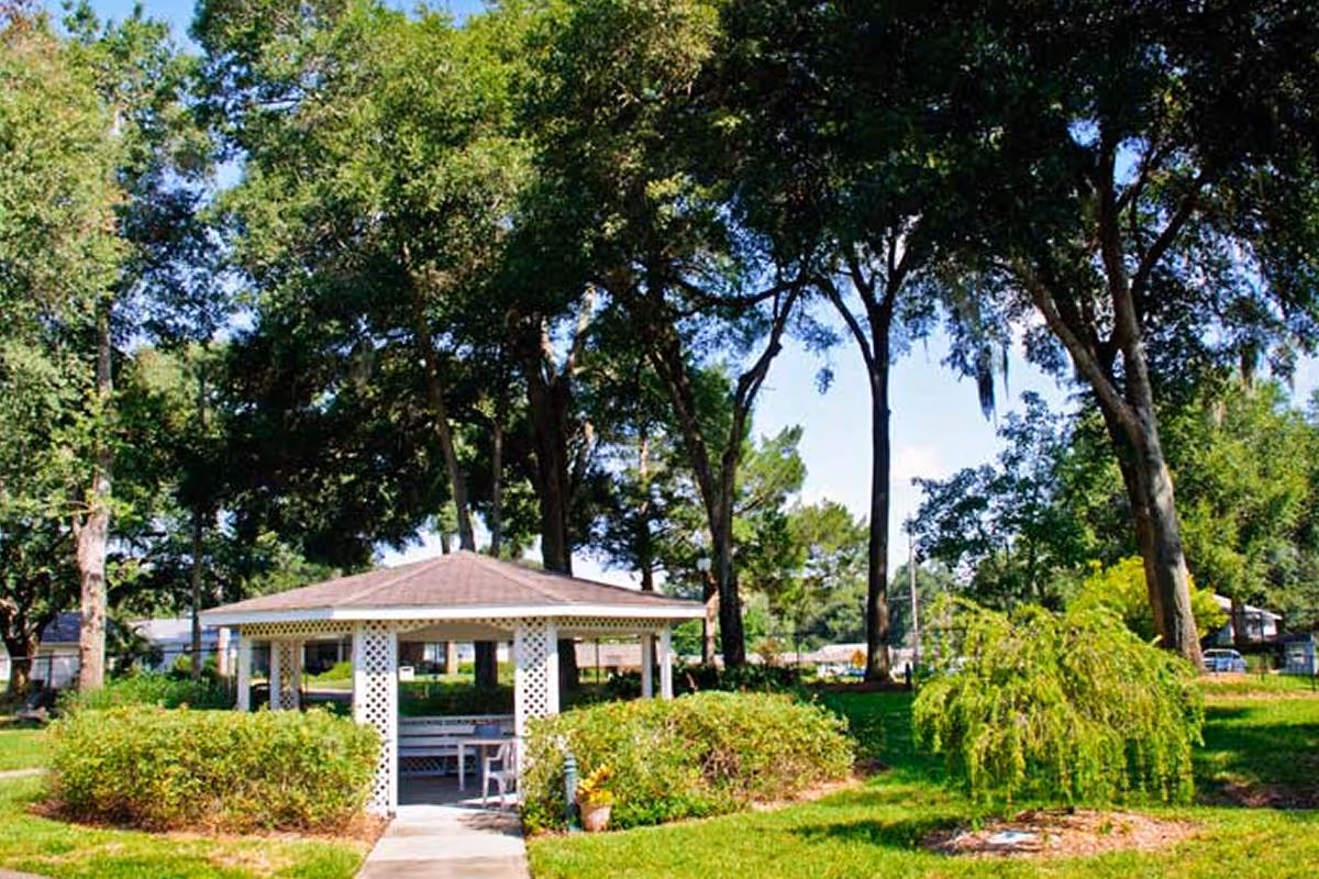 A small white gazebo with lattice sides is situated in a green outdoor area surrounded by trees and bushes under a clear blue sky.