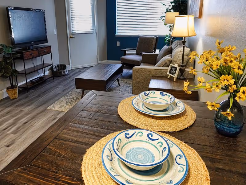 A cozy living room with a dining table set for two in the foreground and a sofa, armchair, coffee table, and TV in the background.
