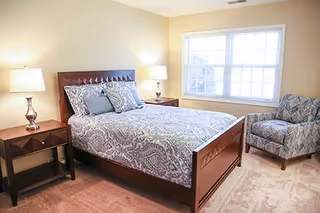A bedroom with a wooden bed frame featuring a patterned gray and white bedspread and pillows. There are two bedside tables with lamps on either side of the bed, a large window letting in natural light, and a patterned armchair in the corner on a carpeted floor.