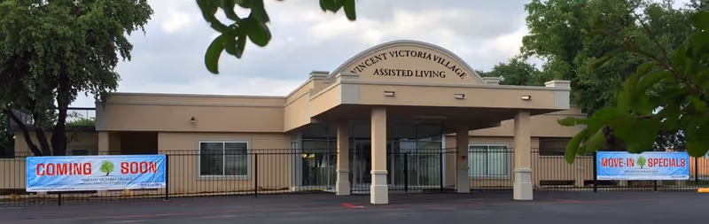Front exterior view of Vincent Victoria Village Assisted Living building with a covered entrance, beige walls, and two banners on the fence reading 'COMING SOON' and 'MOVE-IN SPECIALS'.
