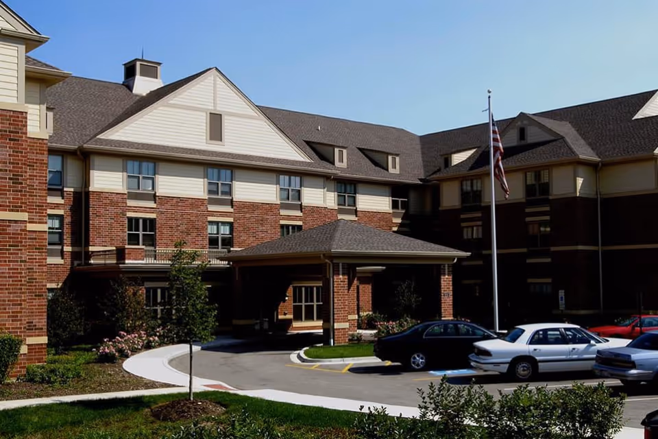 Front entrance of a brick senior living building with a covered porte-cochère, flagpole, landscaping, and parked cars.