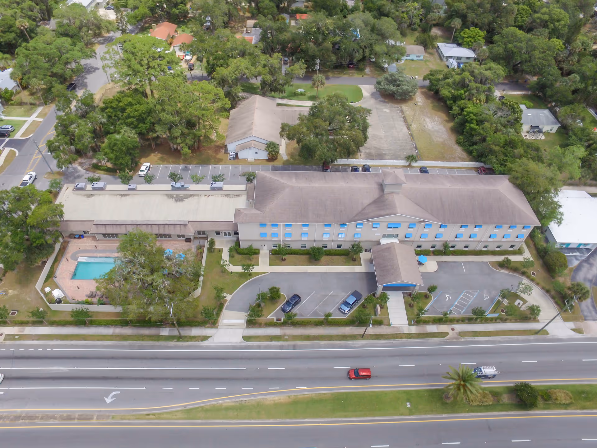 Aerial view of Seashore Senior Living facility showing a large building with blue window awnings, a covered entrance, parking spaces including handicapped spots, a fenced swimming pool area with lounge chairs, and surrounding trees and greenery. The facility is adjacent to a multi-lane road with vehicles driving by.