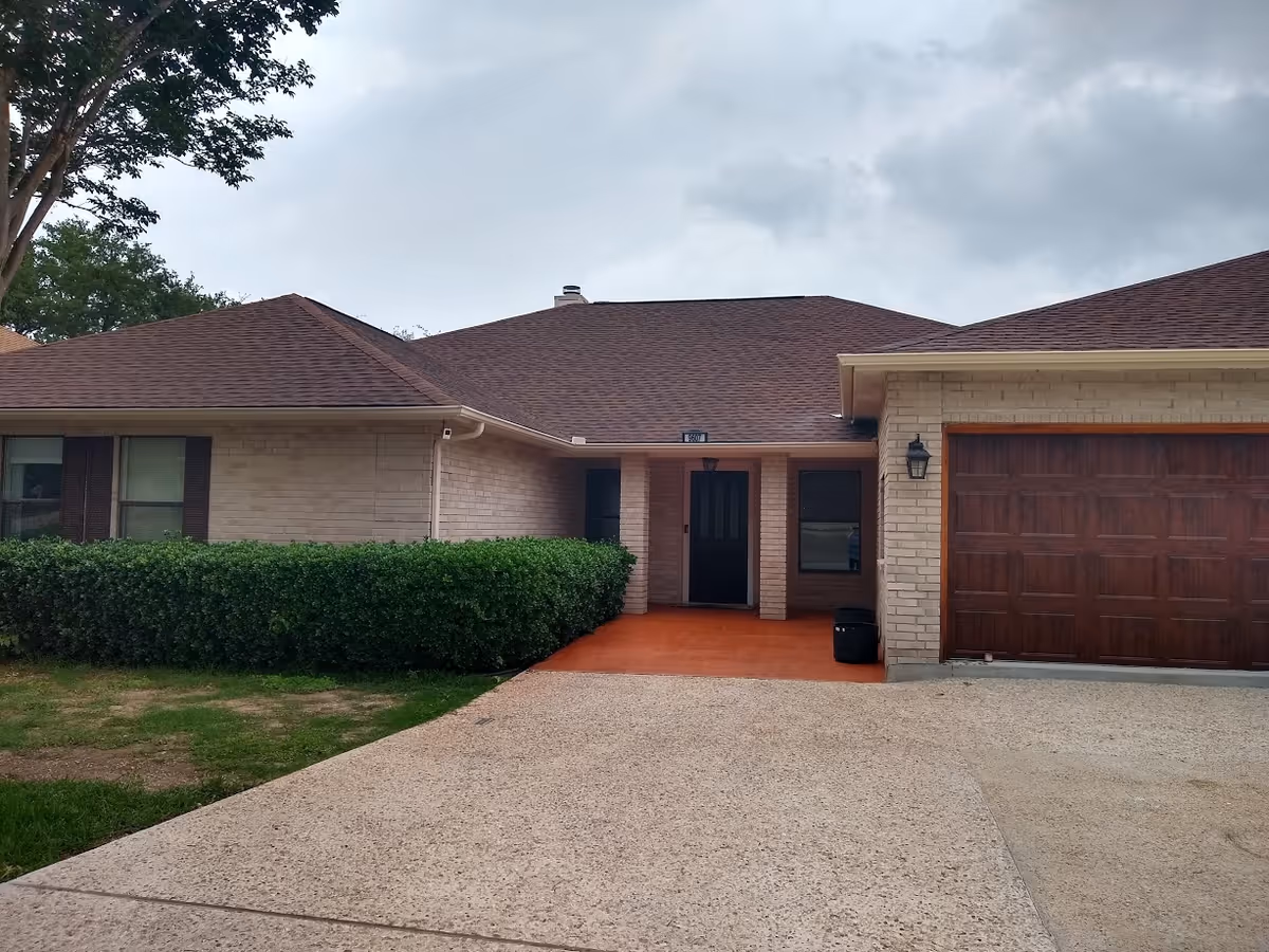 Front exterior view of a single-story brick house with a brown shingled roof, a covered entrance with a black door, two windows, a garage with a wooden door, and a driveway. There are green bushes along the front of the house and a tree on the left side under a cloudy sky.