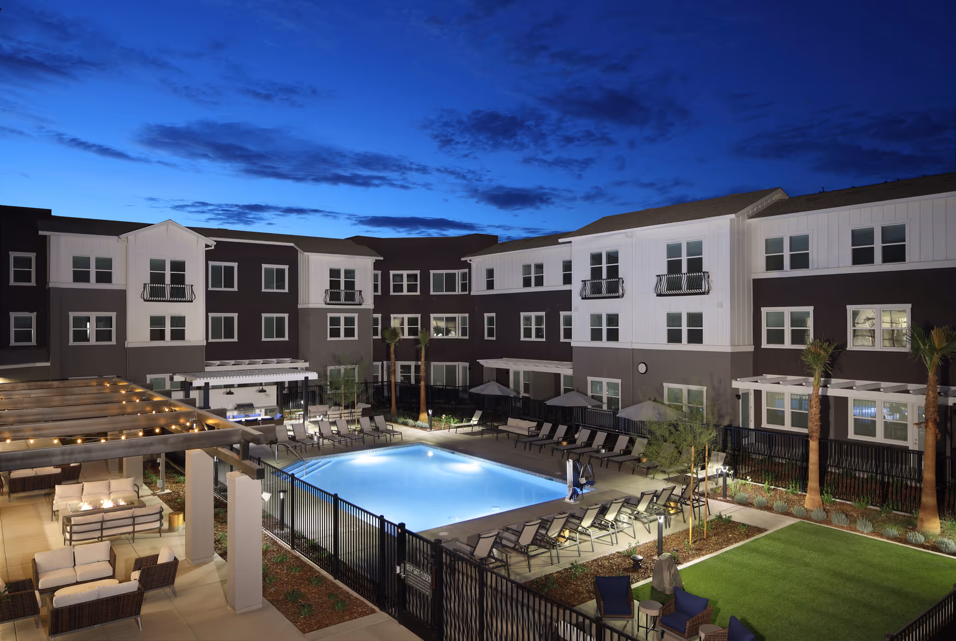Night view of an outdoor courtyard at Sonrisa Senior Living featuring a well-lit swimming pool surrounded by lounge chairs, a pergola with string lights and cushioned seating, palm trees, and a three-story residential building in the background under a partly cloudy sky.