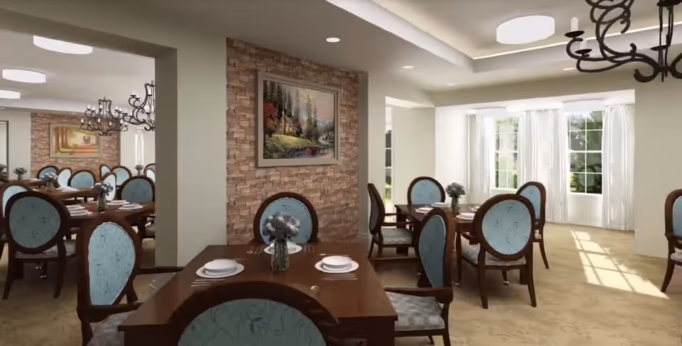 A bright dining room with multiple wooden tables and blue upholstered chairs. Each table is set with white plates, silverware, and a small vase with flowers. The room features large windows with white curtains allowing natural light to fill the space. There is a stone accent wall with a framed landscape painting and elegant chandeliers hanging from the ceiling.