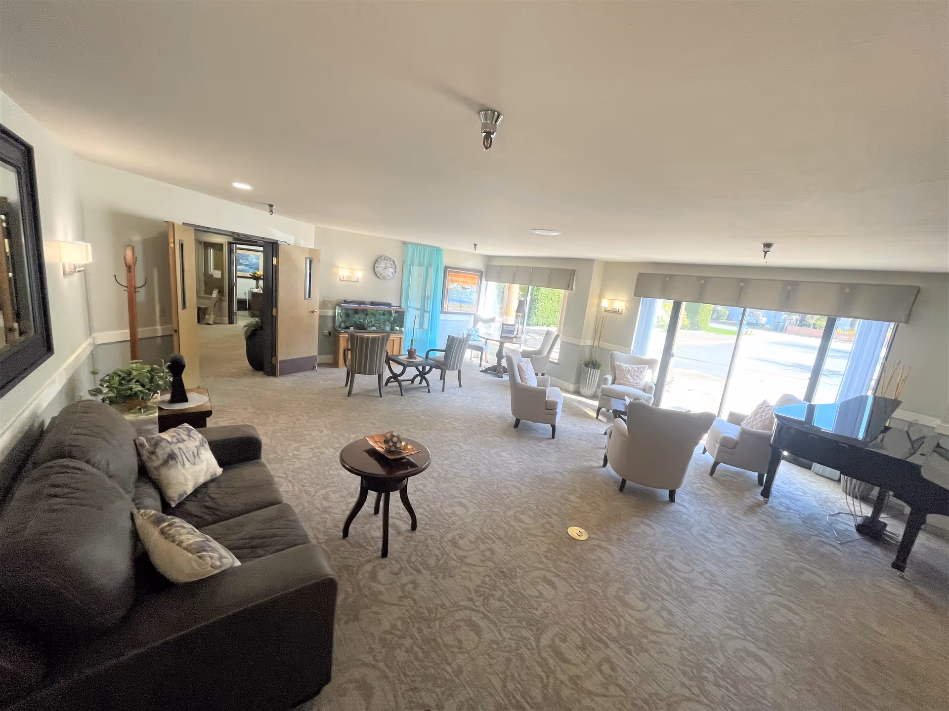 A spacious senior living common area with a dark sofa on the left, several armchairs arranged near large windows letting in natural light, a small round table, a fish tank, and a black grand piano on the right side. The room has light-colored walls and carpet, with wall sconces and a clock visible.