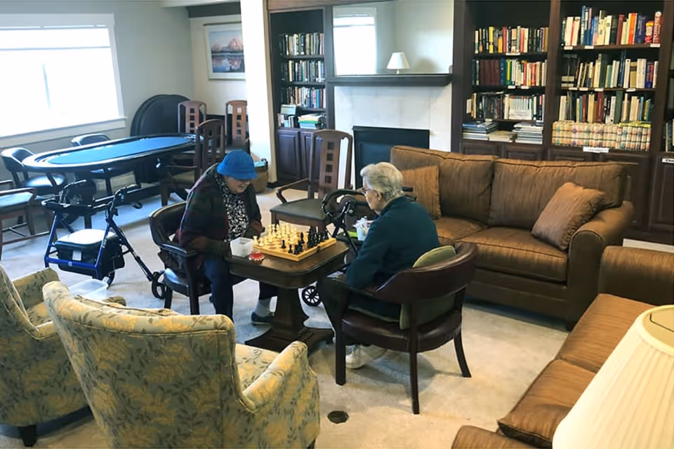 Two individuals playing chess in a cozy lounge with bookshelves and comfortable seating.