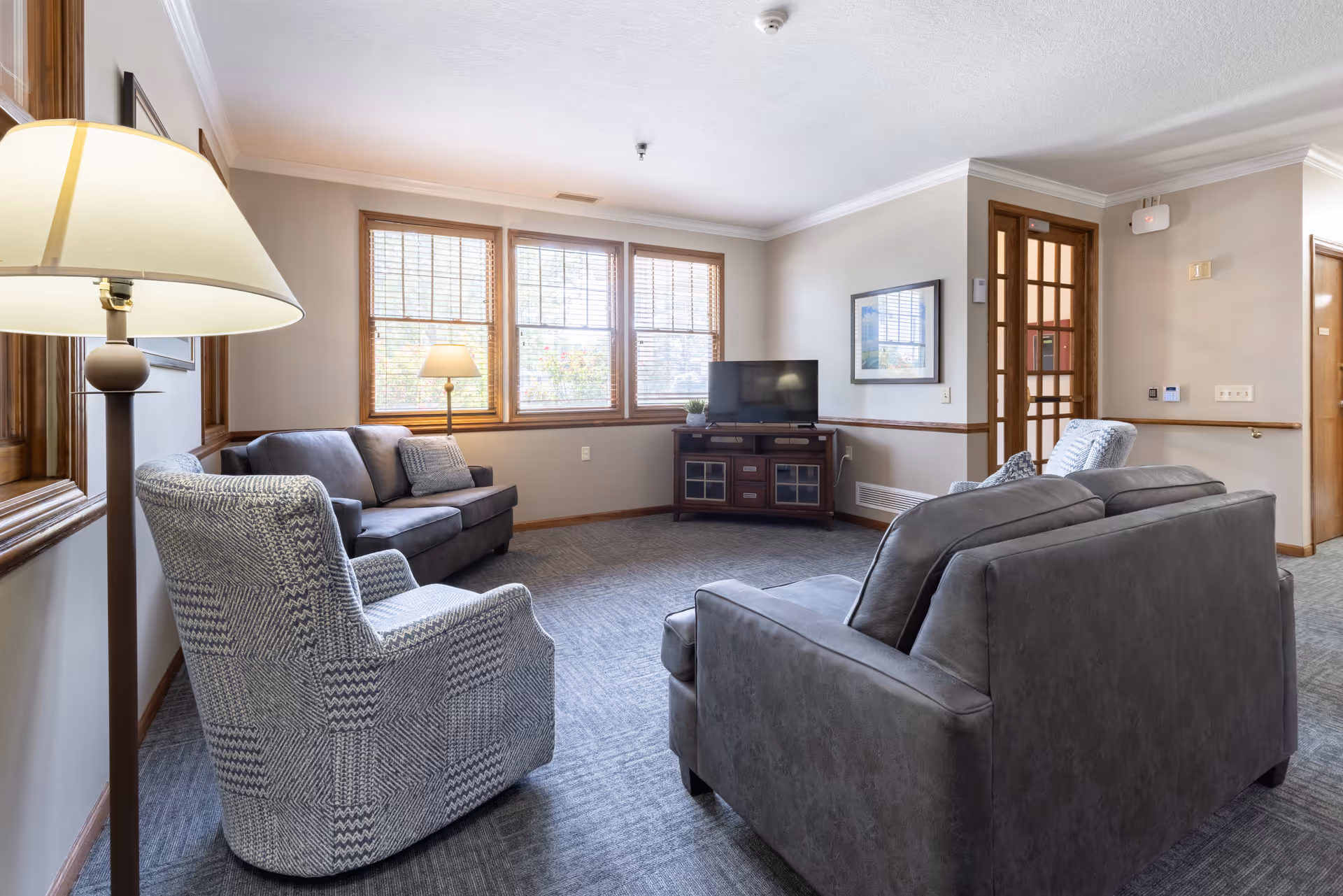 A cozy living room in a senior living facility with two gray sofas, two patterned armchairs, a wooden TV stand with a flat-screen television, and two floor lamps near windows with wooden blinds. The walls are light-colored with wooden trim, and there is a framed picture on one wall.
