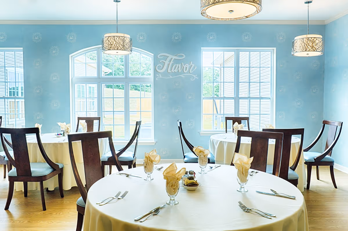 A bright dining room with round tables covered in white tablecloths, each set with folded beige napkins in glasses, silverware, and condiments. The room has light blue walls with large windows letting in natural light and three modern pendant lights hanging from the ceiling. The word 'Flavor' is displayed on the wall between the windows.