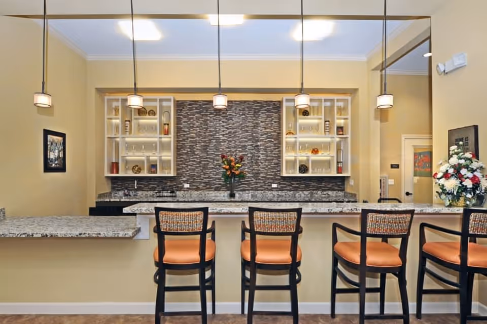 Interior view of a kitchen or bar area with a granite countertop and four high chairs with orange cushions. There are pendant lights hanging from the ceiling, glass shelves with various decorative items, and a flower vase in the center. The walls are painted beige, and there is a framed picture and a flower arrangement on the right side.