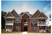 Exterior view of a two-story residential building with brick and beige siding, featuring multiple peaked roofs, balconies, and a central entrance, set against a partly cloudy sky.