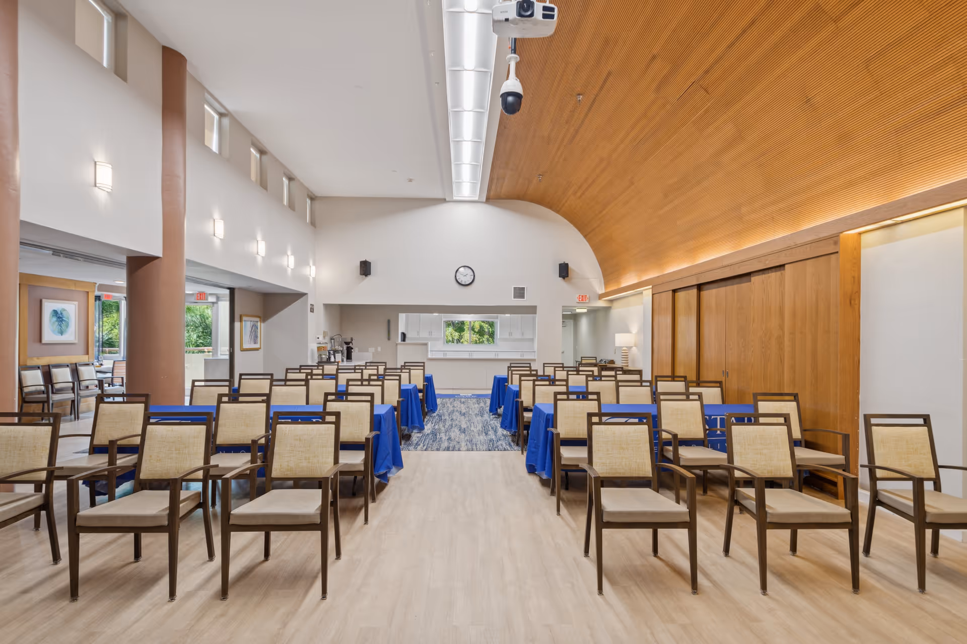 A spacious, well-lit meeting or conference room with rows of wooden chairs with beige cushions arranged facing a front area with tables covered in blue tablecloths. The room features a high ceiling with wood paneling on one side and white walls with small windows near the ceiling. A clock and speakers are mounted on the front wall, and there is a security camera hanging from the ceiling.