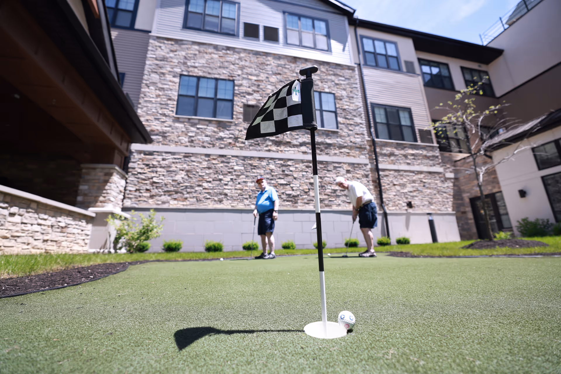 Two elderly men playing putting golf on a small artificial green area in the courtyard of a senior living facility with stone and siding exterior walls in the background.