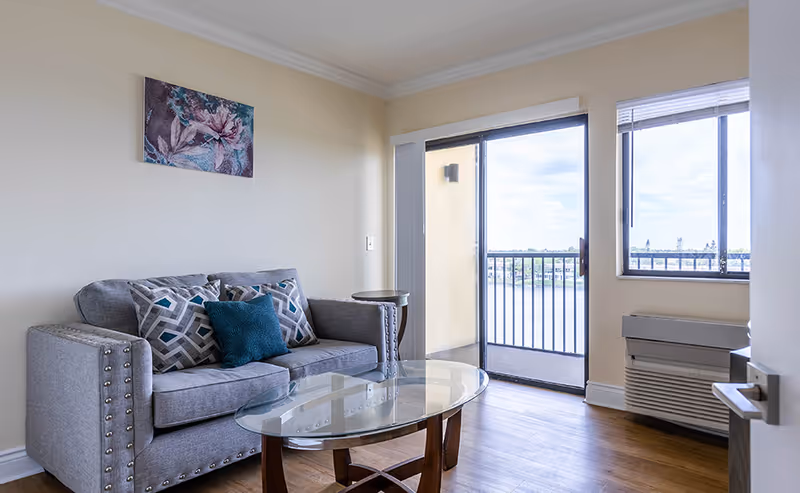 A small living room with a gray couch adorned with three decorative pillows, a glass coffee table with wooden legs, a small round side table, and a floral painting on the wall. There is a sliding glass door leading to a balcony overlooking a body of water, and a window with blinds next to an air conditioning unit.