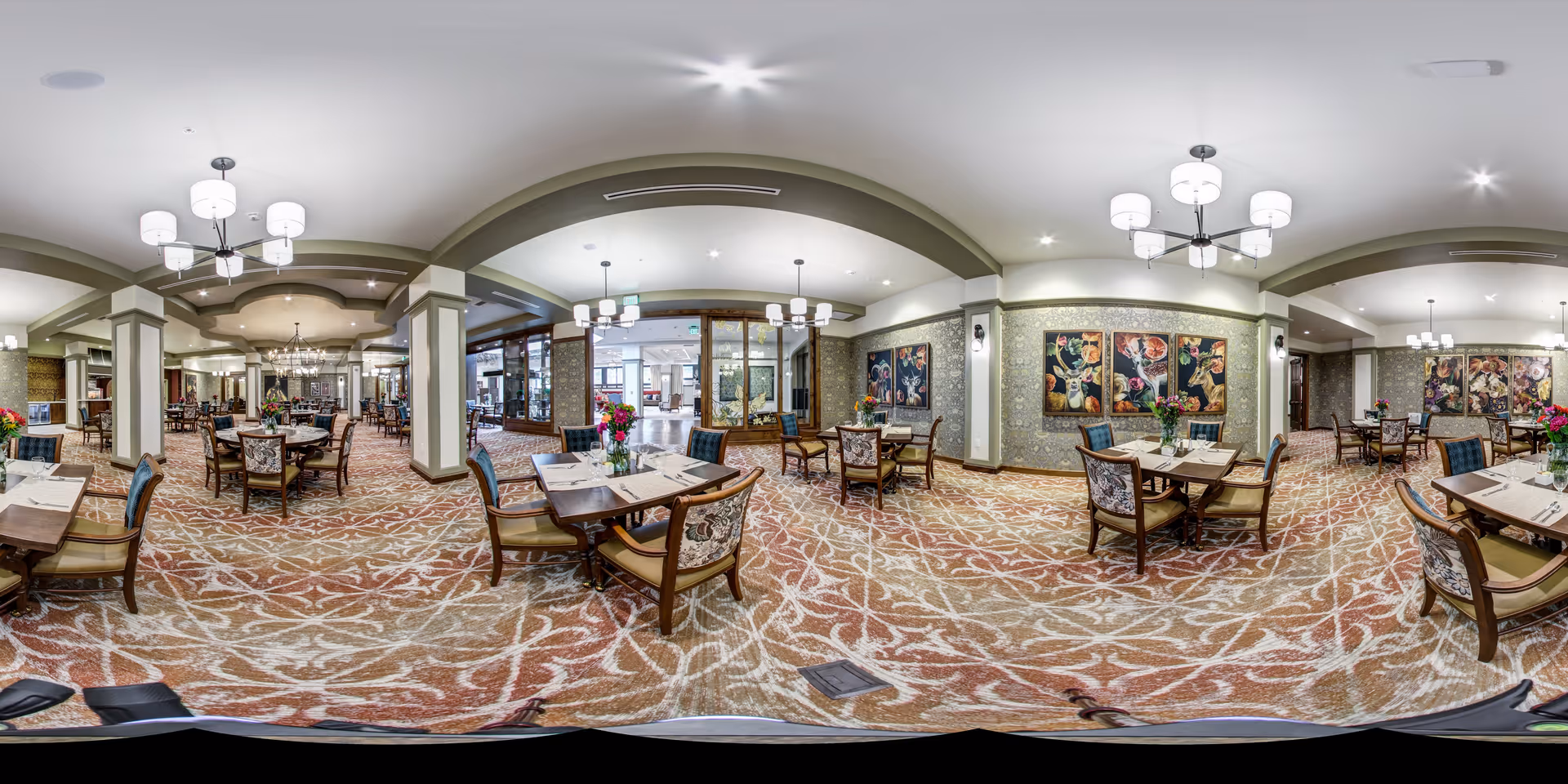 Spacious dining room with multiple set tables and upholstered chairs, chandeliers, floral artwork, and a patterned carpet.