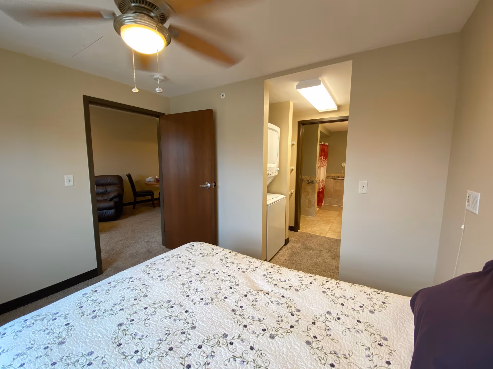 View from a bedroom showing a bed with a floral quilt, an open door leading to a living area with a recliner and chairs, a stacked washer and dryer unit in a small laundry nook, and a bathroom with a red shower curtain in the background.