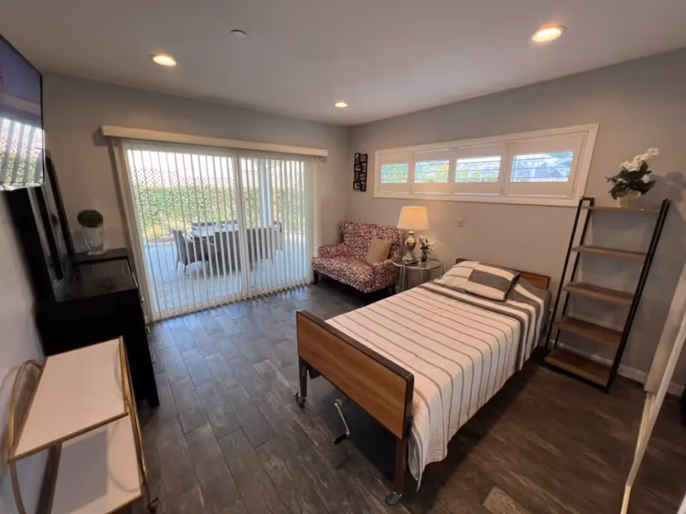 A well-lit bedroom with a single bed covered in a striped blanket, a patterned loveseat, a side table with a lamp and flowers, a shelving unit with a plant, and sliding glass doors leading to an outdoor patio with a table and chairs. The room has wood-look flooring and small windows near the ceiling.