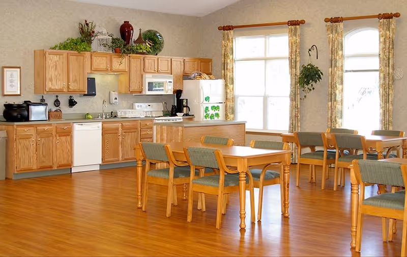 A bright dining area with wooden tables and green cushioned chairs. The room features a kitchen area with wooden cabinets, a white dishwasher, stove, microwave, and refrigerator. Large windows with floral curtains allow natural light to fill the space. There are decorative plants and vases on top of the cabinets and a hanging plant near the window.