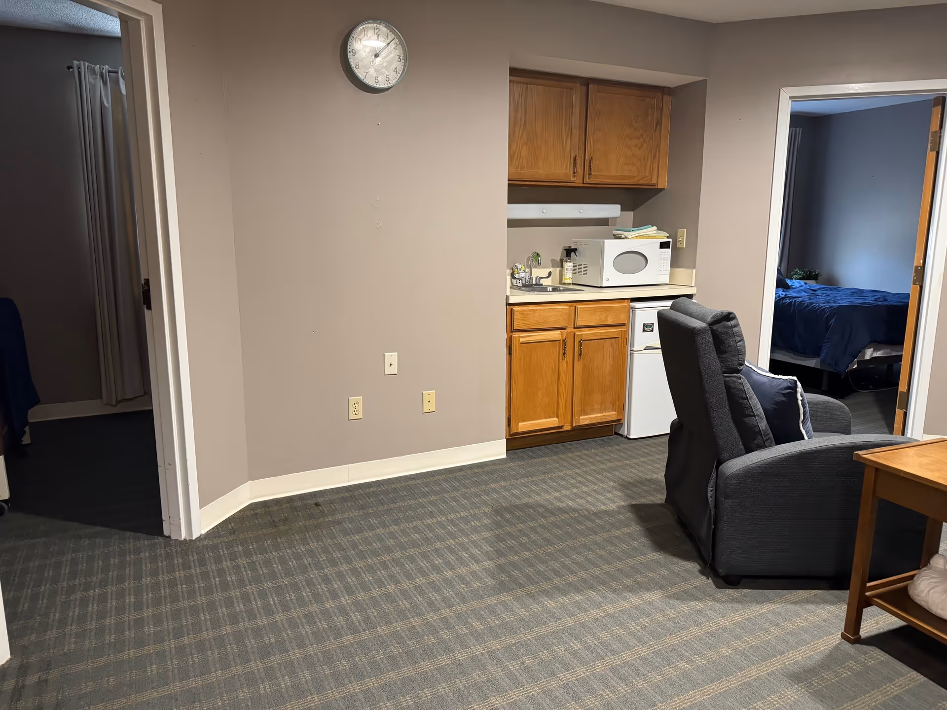 Interior view of a senior living facility room showing a small kitchenette with wooden cabinets, a microwave, and a mini refrigerator. A gray recliner chair with a pillow is positioned near the kitchenette. Two doorways lead to bedrooms with beds visible inside. A wall clock is mounted above the kitchenette area.