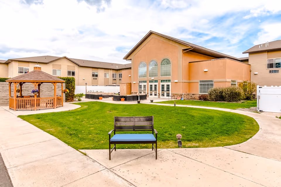 Outdoor courtyard area of Ellensburg Senior Living featuring a green lawn, a wooden gazebo with hanging flower pots, a bench with a blue cushion, and a large building with tall arched windows in the background under a partly cloudy sky.