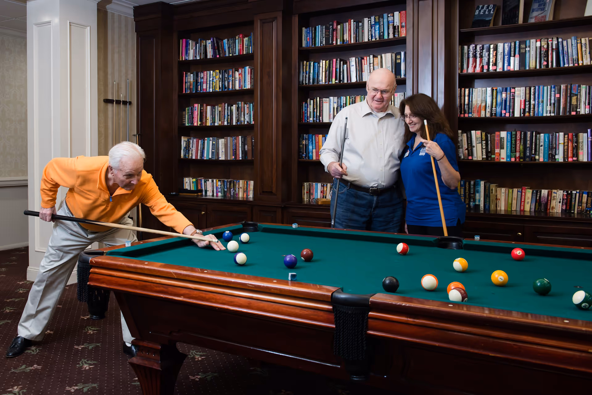 Three seniors playing pool in a room with dark wooden bookshelves filled with books. One man in an orange sweater is taking a shot while another man and a woman stand nearby holding pool cues and smiling.