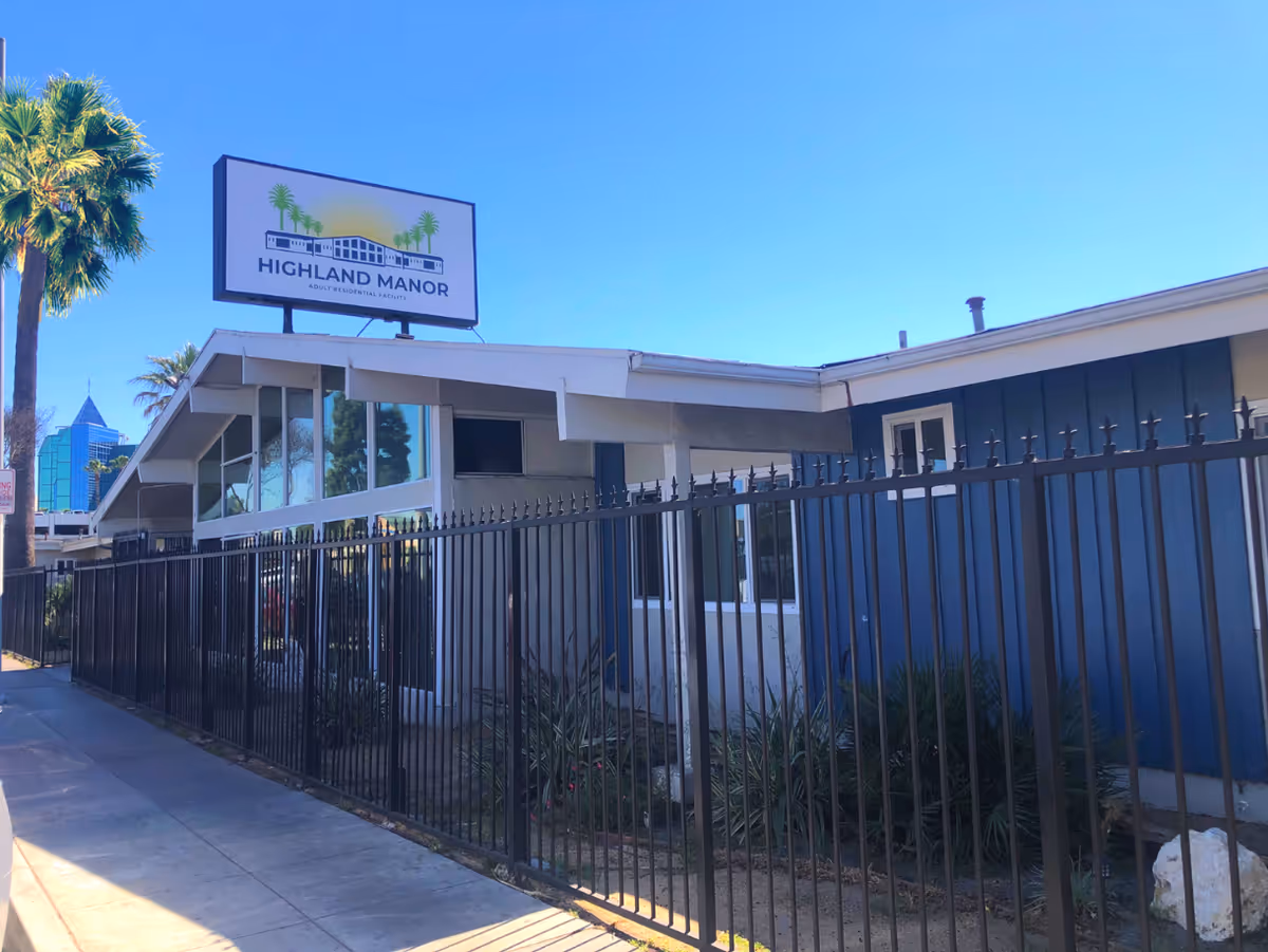 Exterior front of Highland Manor assisted living building with a sign, black fence, and palm trees under a clear blue sky.