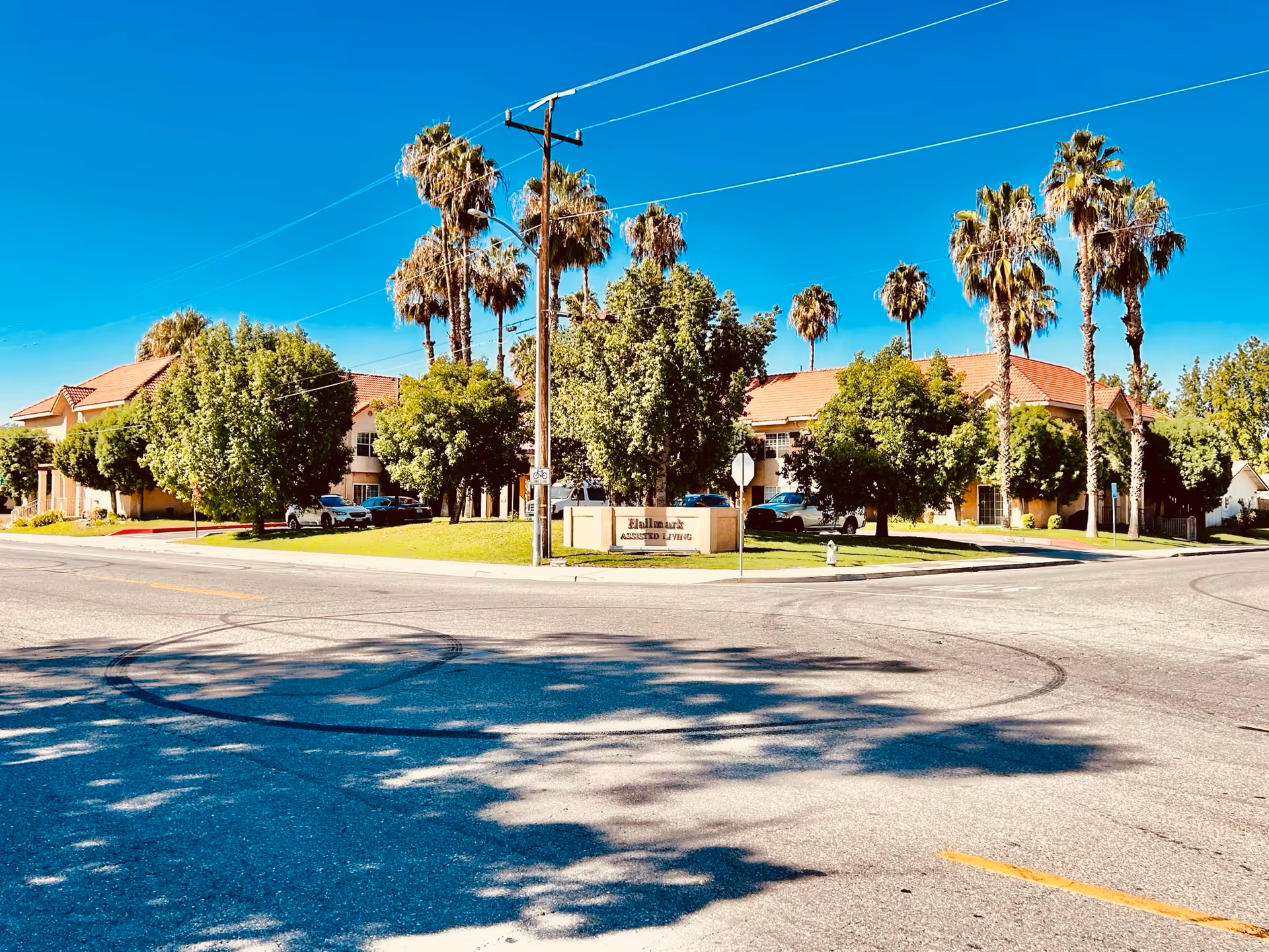 Exterior view of Hallmark of Bakersfield assisted living facility with a clear blue sky, palm trees, and other green trees surrounding the building. The building has a red-tiled roof and beige walls. A sign in front reads 'Hallmark Assisted Living'.