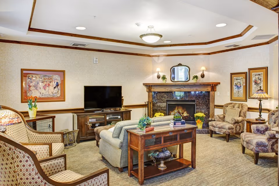 A cozy living room in a senior living facility featuring a fireplace with a lit fire, a flat-screen TV on a wooden cabinet, a sofa, patterned armchairs, a wooden side table with books and plants, framed artwork on the walls, and warm lighting from wall sconces and a ceiling light.