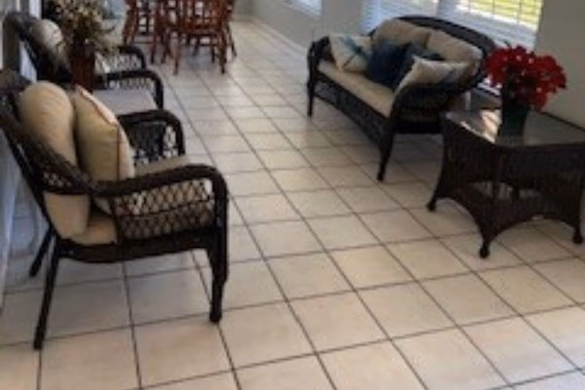 Sunlit seating area with wicker chairs and a loveseat on a tiled floor, a side table with flowers, and a dining table visible in the background.