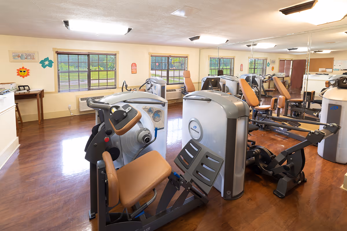 A well-lit exercise room with several pieces of Nautilus fitness equipment arranged on a polished wooden floor. The room has large windows with blinds, a mirrored wall reflecting the equipment, and light-colored walls decorated with colorful wall art.