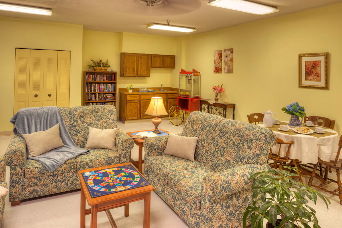 Bright communal lounge with patterned sofas and pillows, a small game table, kitchenette and dining table set.