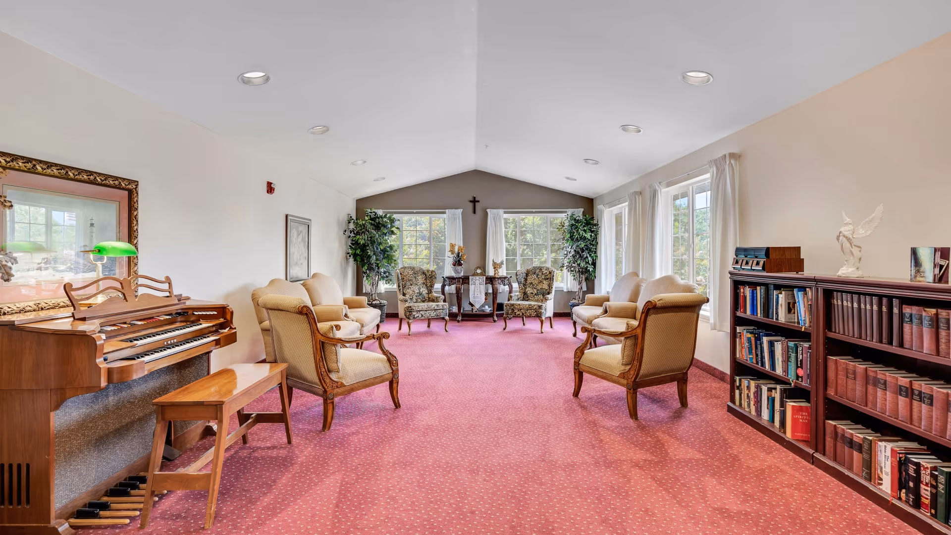A spacious living room with a red carpet and cream-colored walls. The room features two beige sofas, four armchairs with floral upholstery, a wooden organ with a green lamp, and a wooden bookshelf filled with books. Large windows with white curtains allow natural light to fill the room. A small table with decorative items and plants is positioned between the windows, and a cross hangs on the far wall.