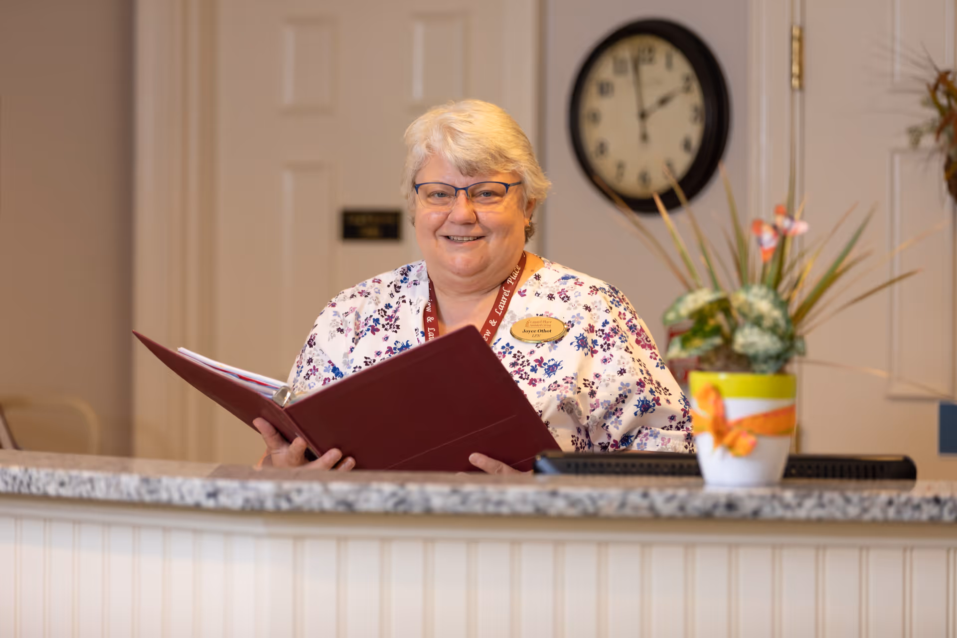 Reception area inside an assisted living facility with a smiling staff member standing behind the front desk holding a red binder, a clock on the wall and a potted plant on the counter.