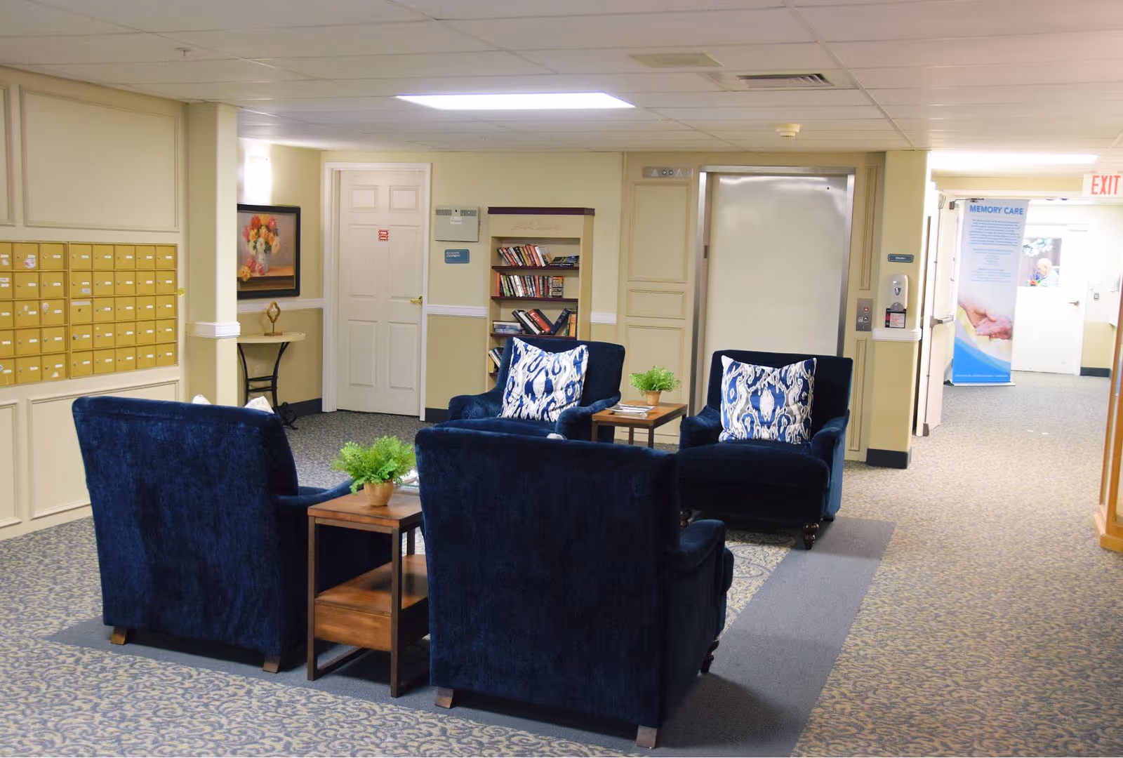 A seating area in a senior living facility with four dark blue armchairs arranged around two small wooden tables with potted plants. The area is carpeted with patterned flooring and is located in a hallway with mailboxes on the left wall, a bookshelf, and an elevator door in the background. There is a sign indicating 'Memory Care' visible down the hallway.
