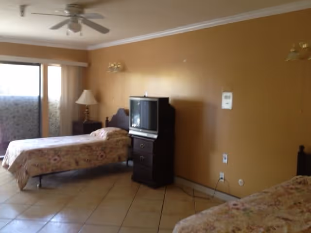 A senior living facility bedroom with two twin beds covered in floral bedspreads. There is a wooden nightstand with a lamp between the beds and a small TV on a dark wooden dresser against a tan wall. The room has tiled floors, a ceiling fan, and a sliding glass door with curtains leading to an outdoor area.