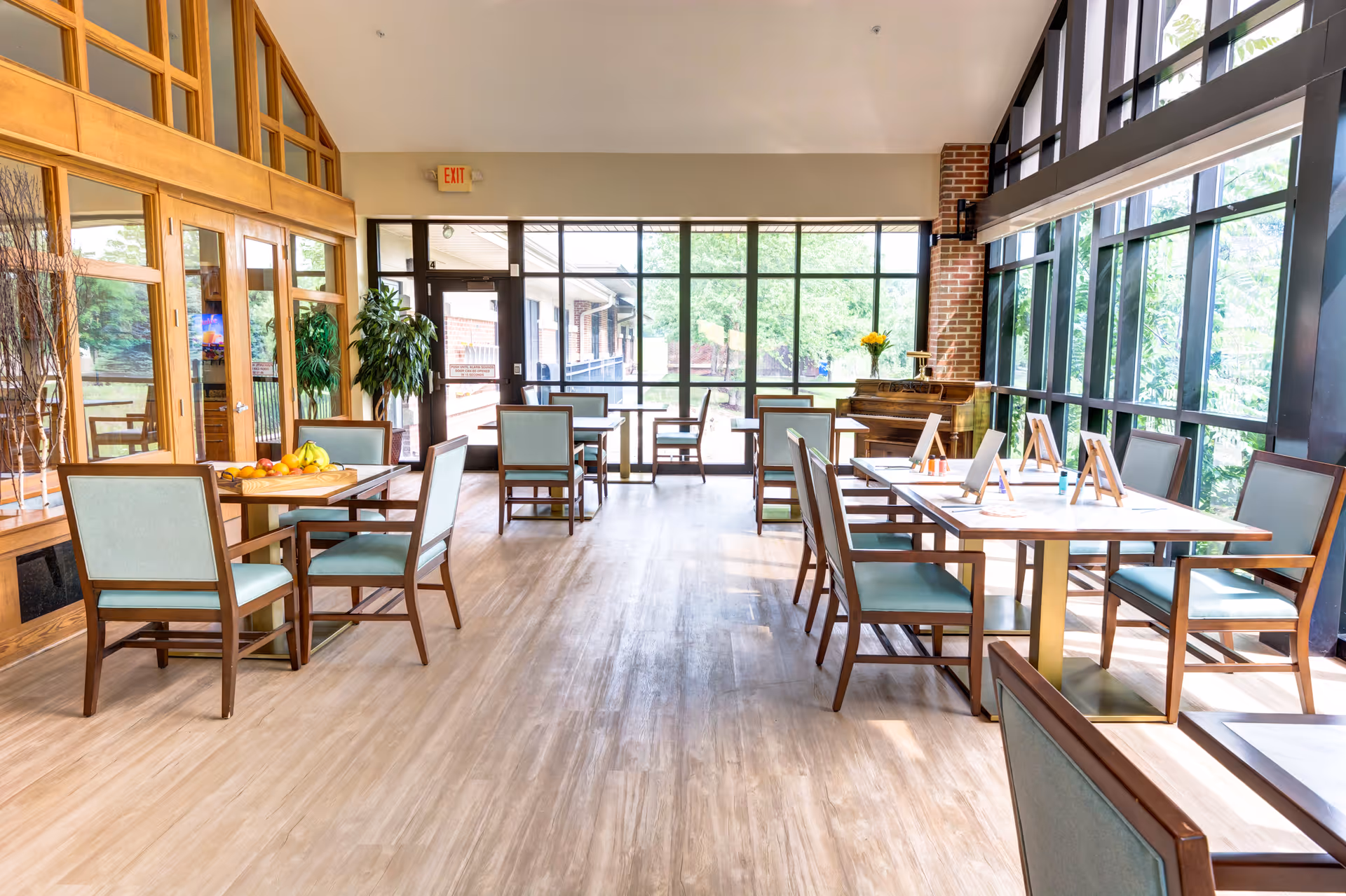 Bright and spacious common area with large windows letting in natural light, several tables and chairs arranged for seating, a piano in the corner, and a bowl of fruit on one table. The room has wooden floors and a mix of brick and glass walls.