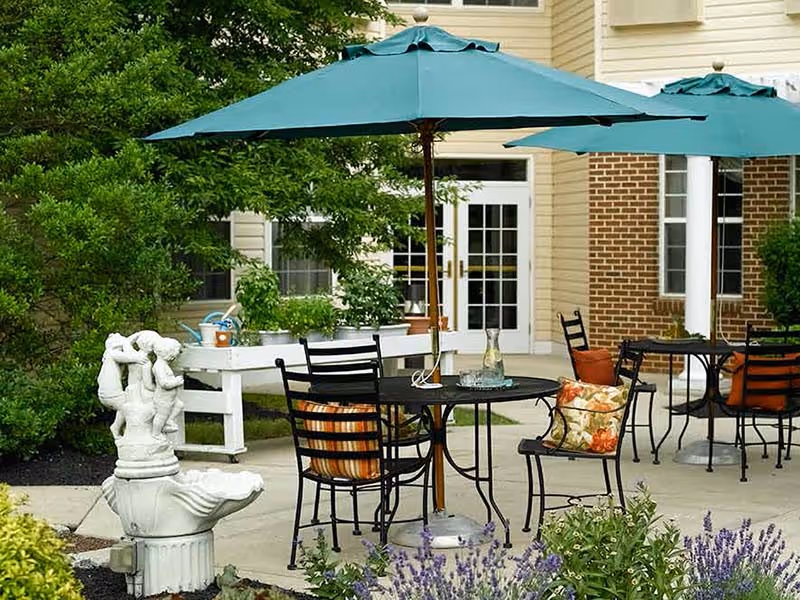 Outdoor patio area with black metal tables and chairs, each table shaded by a large green umbrella. The chairs have colorful cushions with floral and striped patterns. There is a white decorative fountain sculpture in the foreground and various plants and flowers surrounding the patio. The background shows a building with beige siding and brick walls, and white-framed glass doors.
