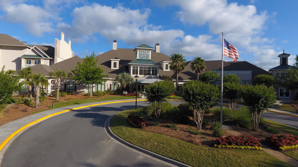 Front entrance of a senior living facility with a circular driveway, landscaped islands, palm trees, and an American flag.