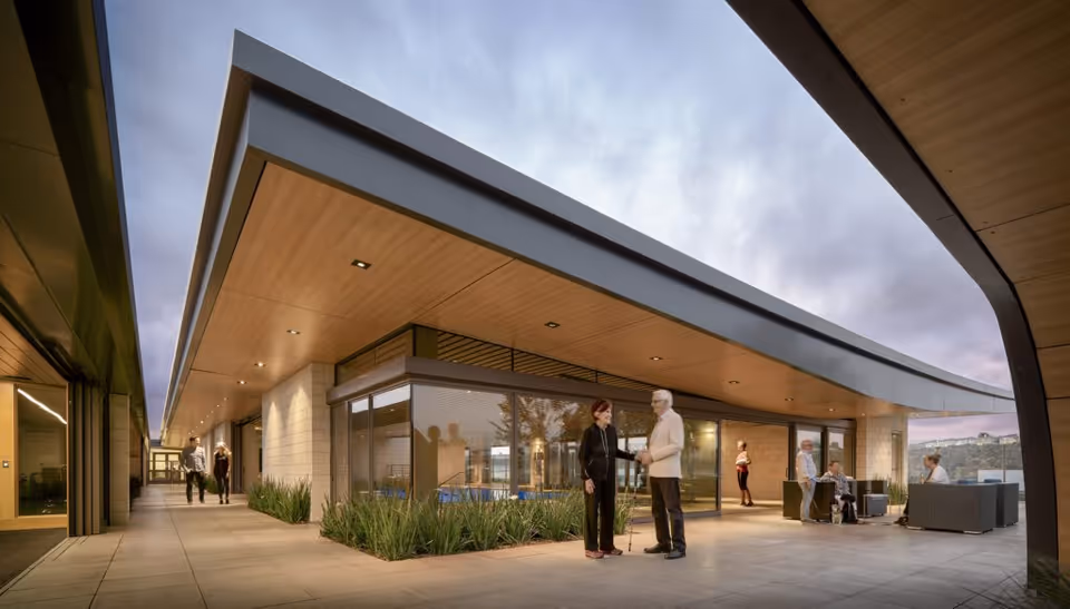 Modern senior living facility courtyard with a covered walkway and residents conversing on a tiled terrace at dusk.