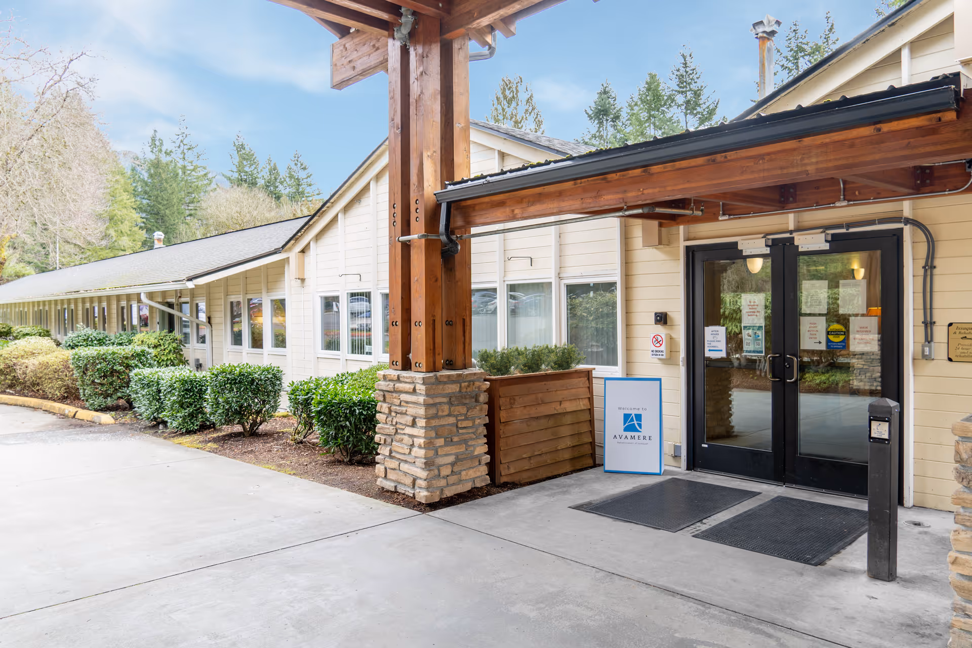 Entrance of Avamere Rehabilitation of Issaquah facility showing double glass doors under a wooden canopy with stone pillars, surrounded by bushes and trees.